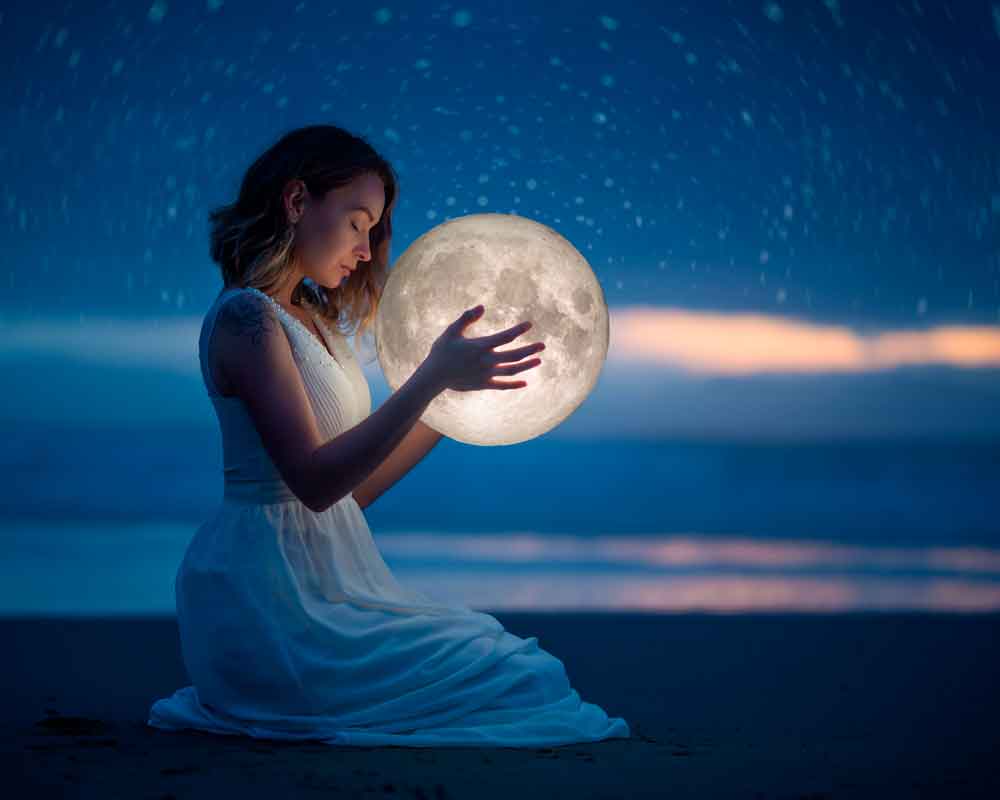 Mulher sentada na praia, na beira do mar, à noite, segurando a lua nas mãos