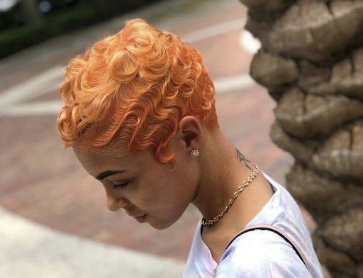 Jovem mulher negra com colar dourado no pescoço e finger waves em seu cabelo laranja.