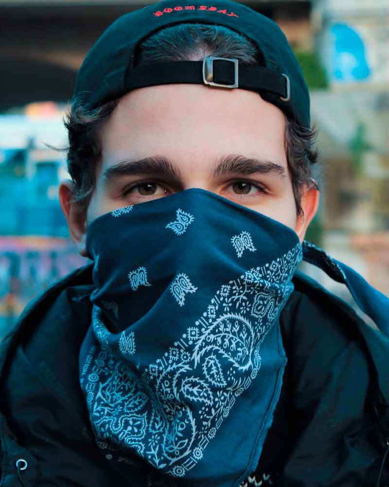 Homem jovem na rua com boné para trás, jaqueta de frio e bandana azul no rosto como máscara para prevenção do coronavírus, estilo na pandemia