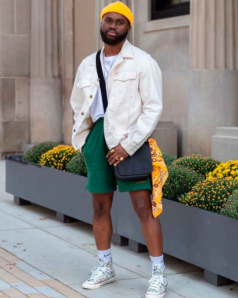 Homem jovem negro cheio de estilo com converse branco, shorts verde, jaqueta jeans creme, touca amarela, bandana amarela e bolsa preta tendência streetstyle