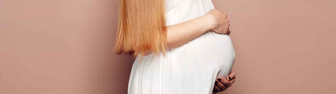 Mulher jovem grávida de cabelos longos, volumosos, lisos e loiros, segurando a barriga com as mãos em fundo neutro nude bege