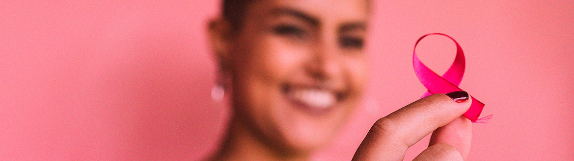 Uma mulher com o cabelo raspado, em segundo plano, segura na mão um laço rosa, que aparece em primeiro plano.